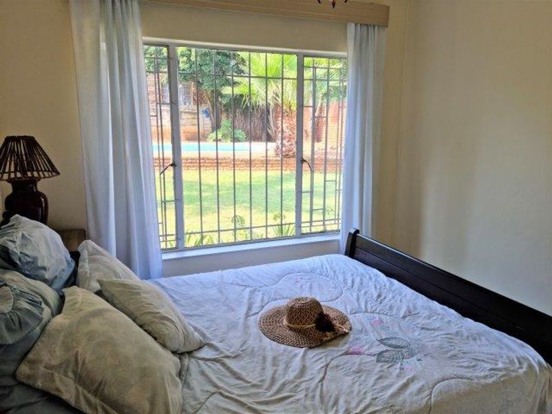 A bedroom featuring a large window with security bars, offering a view of the garden and pool area.