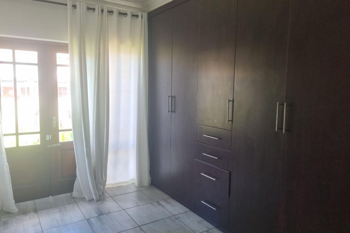 A bright bedroom featuring dark wood built-in cupboards, tiled flooring, and direct access to the exterior through a timber-framed glass door.