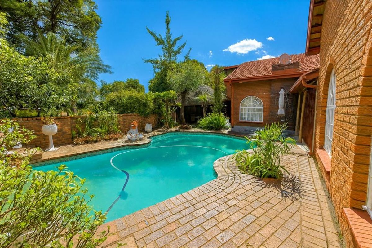 A well-maintained outdoor pool area featuring brick paving, a surrounding brick boundary wall, and a charming thatched lapa in the garden.