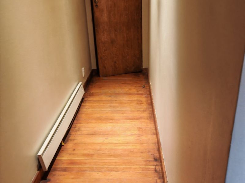 A narrow hallway featuring original hardwood flooring and a built-in baseboard heater, leading to a wooden interior door.