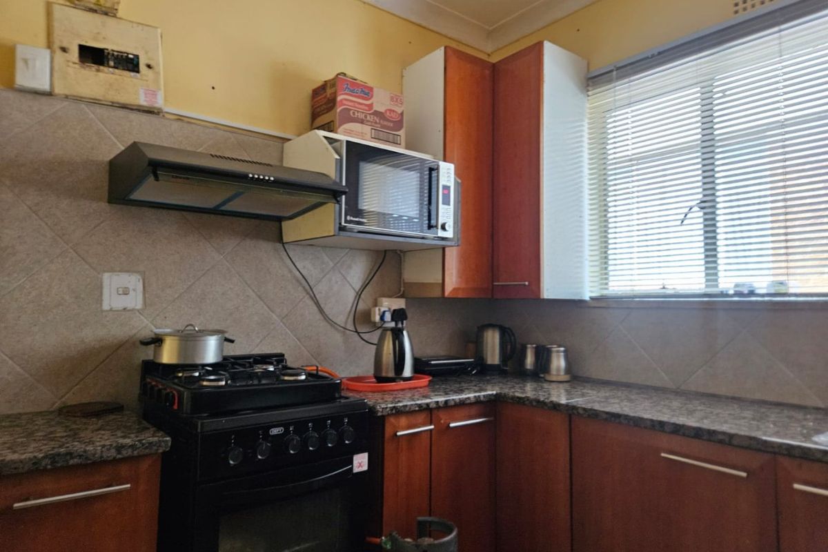 A compact kitchen featuring wooden cabinetry, granite countertops, a gas stove with extractor fan, and tiled backsplash, requiring cosmetic updates.
