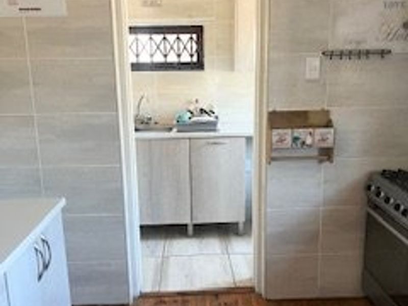 A kitchen area featuring timber flooring, tiled walls, and built-in cabinetry, with an adjacent scullery or prep area visible through the doorway.