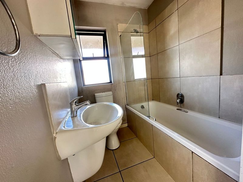 A functional bathroom featuring a pedestal basin, a bathtub with a glass shower screen, and neutral wall and floor tiling.
