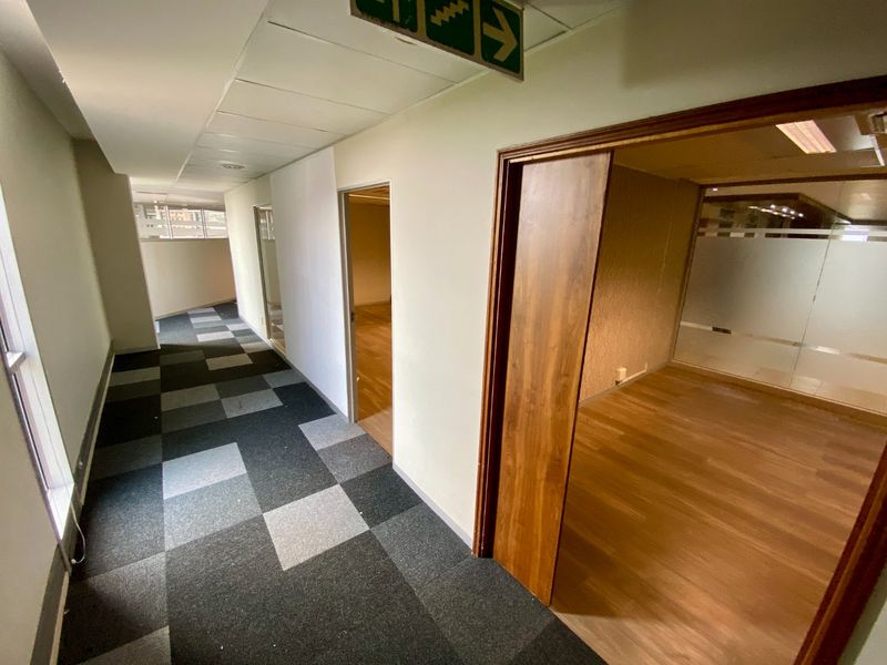 A commercial office hallway featuring a mix of carpet tile and laminate flooring, leading into vacant office spaces with glass partitions.