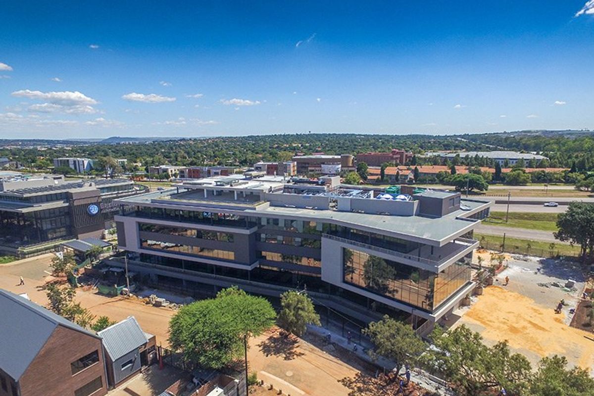 Aerial view of a modern commercial office building featuring a glass facade and rooftop infrastructure, situated within a developing business park environment.