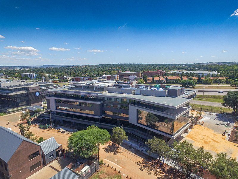 Aerial view of a modern commercial office building featuring a glass facade and rooftop infrastructure, situated within a developing business park environment.