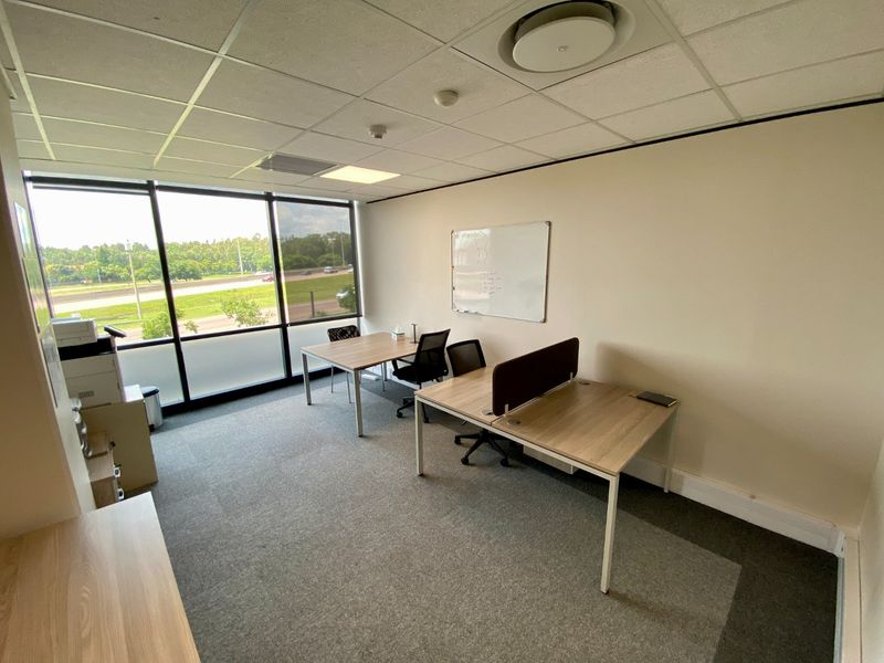 A bright, functional office space featuring large windows with natural light, carpeted flooring, and a suspended ceiling.