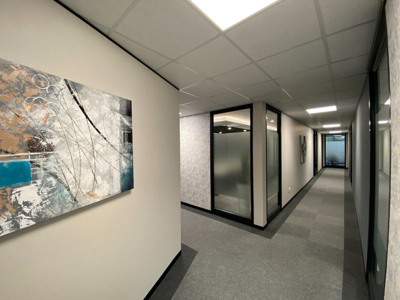 A modern commercial office hallway featuring carpeted flooring, suspended ceiling with recessed lighting, and glass-partitioned office spaces.