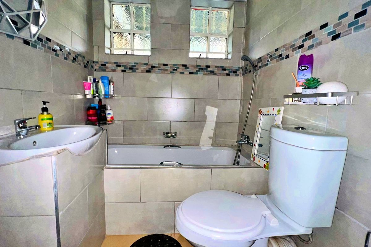 A compact bathroom featuring a built-in bathtub, pedestal washbasin, and toilet, finished with neutral wall tiles and a decorative mosaic border.