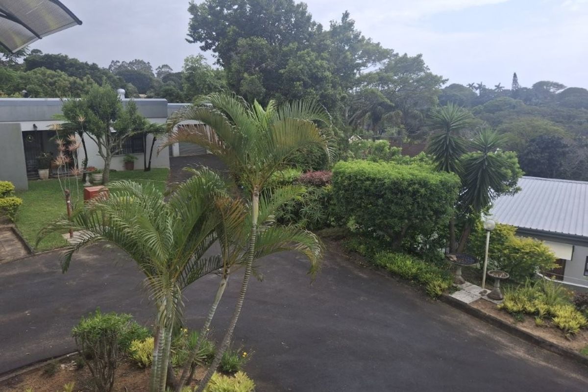 An elevated view of the rear property grounds featuring a paved driveway, established garden landscaping, and mature trees surrounding the townhouse exterior.