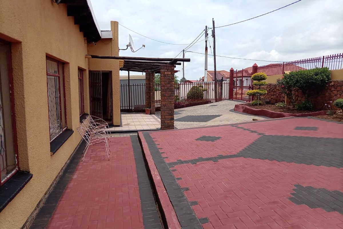 Rear exterior view featuring a paved driveway, brick pergola structure, and secure boundary walling with metal fencing.