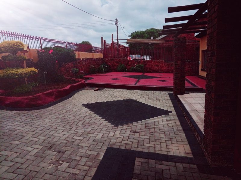 A paved outdoor patio area featuring a timber pergola, brick retaining walls, and decorative patterned paving, enclosed by metal perimeter fencing.