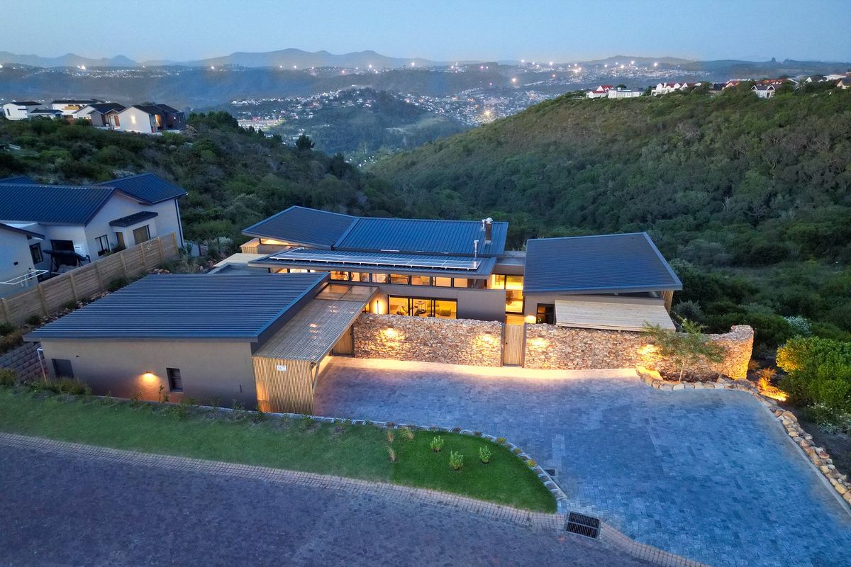 Aerial view of a modern residence featuring stone cladding, solar panels, and a paved driveway, set against a scenic hillside landscape at dusk.