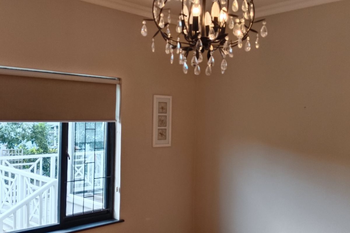 A bright bedroom featuring a decorative chandelier, neutral wall tones, and a window with a roller blind overlooking an outdoor balcony area.