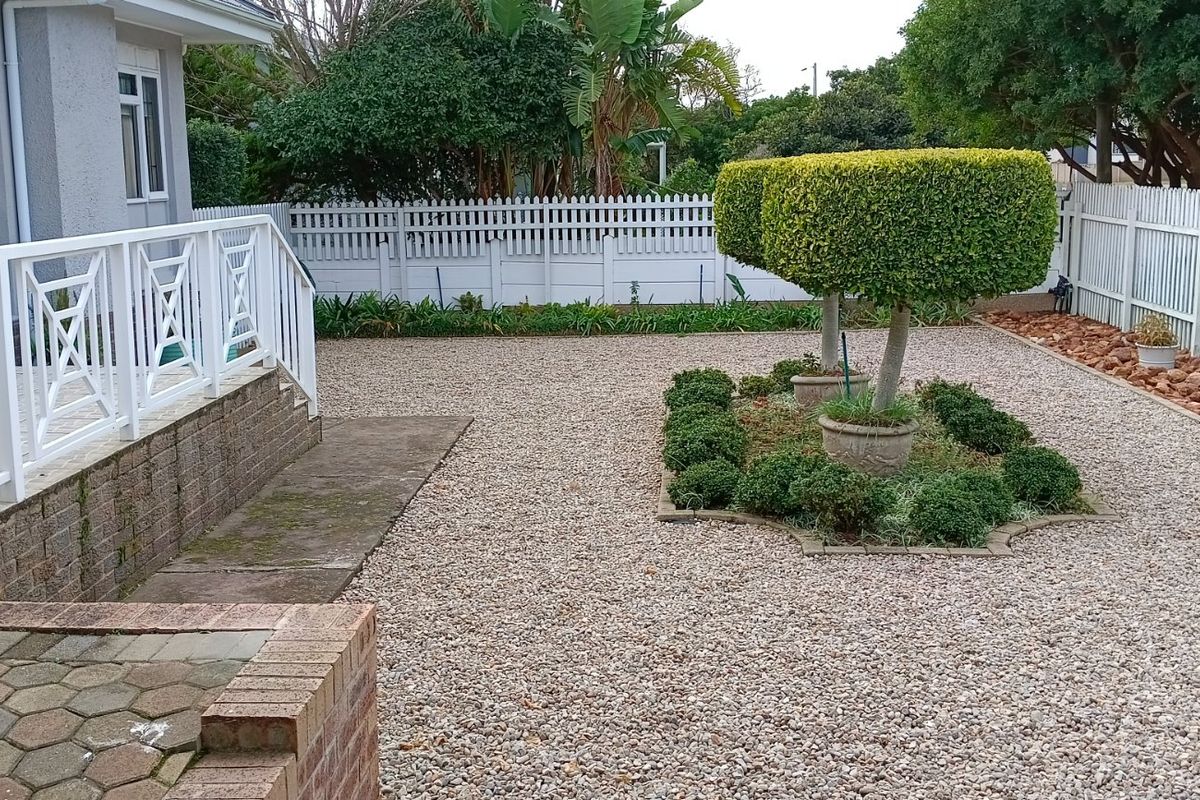 A well-maintained rear garden featuring a gravel landscape, manicured topiary trees, a white picket fence, and a paved patio area with white railings.