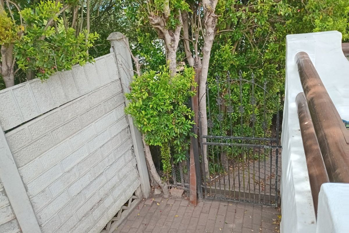 A rear exterior area featuring a paved walkway, a wrought iron gate, and a leaning precast concrete boundary wall with mature surrounding trees.