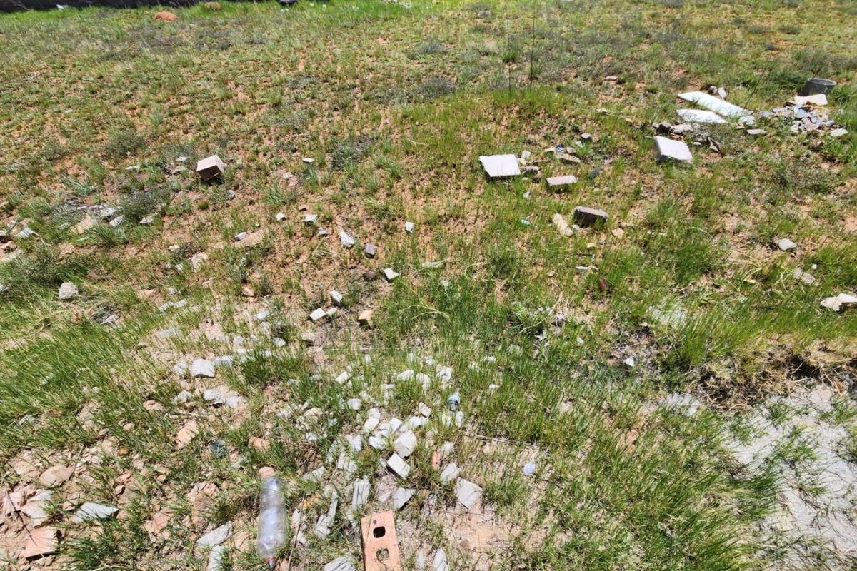 A view of a vacant residential plot featuring natural grass cover and a perimeter boundary wall, currently containing scattered construction debris.