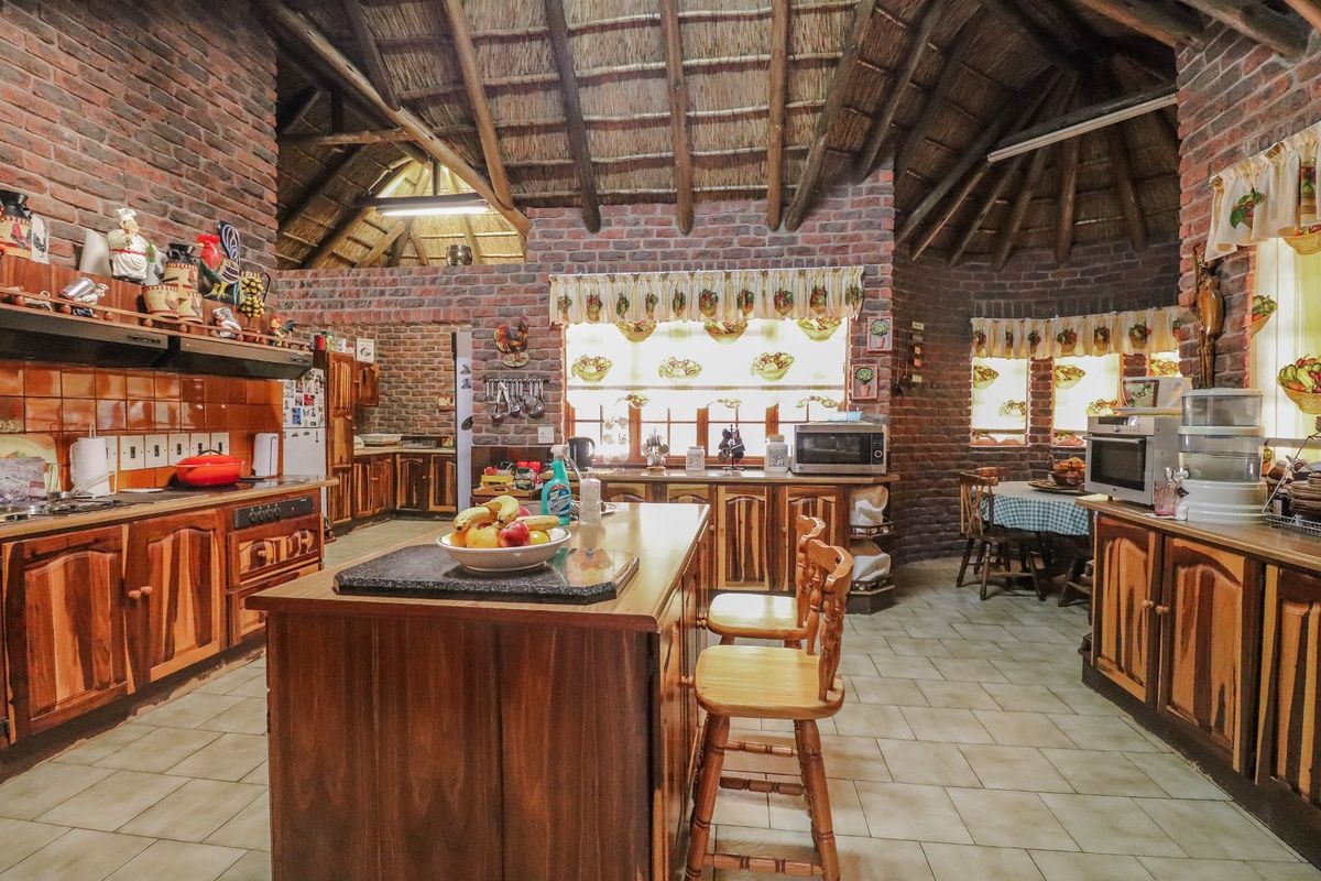 Spacious rustic kitchen featuring exposed brick walls, a high thatched ceiling, timber cabinetry, and a central kitchen island with tiled flooring.