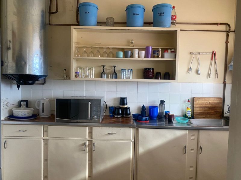 A dated kitchen featuring original cabinetry, exposed copper piping, a stainless steel sink, and tiled splashback, offering significant potential for modernization.