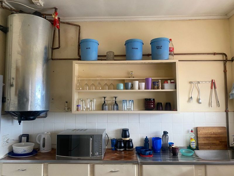 A dated kitchen area featuring exposed copper plumbing, a wall-mounted water heater, tiled splashback, and basic cabinetry with a stainless steel sink.
