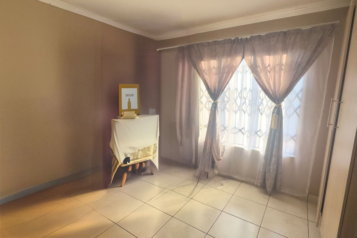A simple, neutrally painted bedroom featuring tiled flooring, a security gate on the window, and a built-in cupboard.