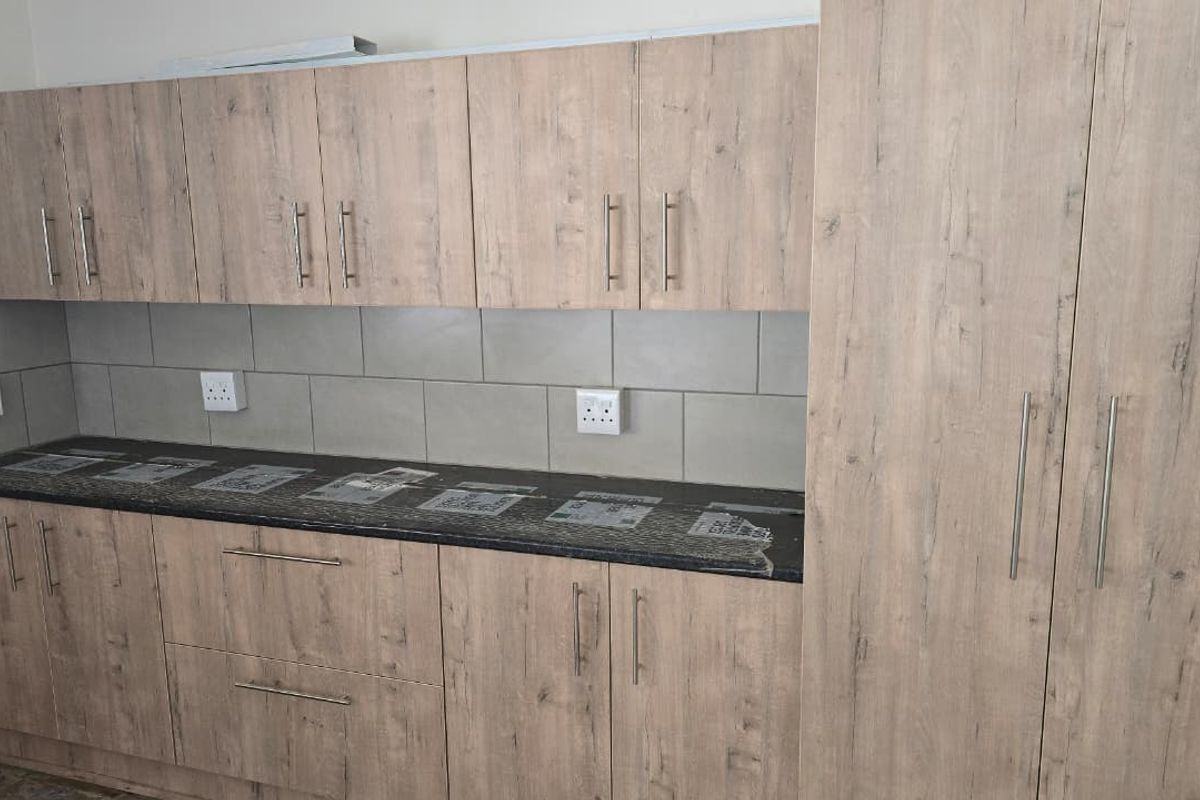 A modern kitchen area featuring light wood-grain laminate cabinetry, dark stone countertops, and a neutral tiled backsplash with ample storage space.