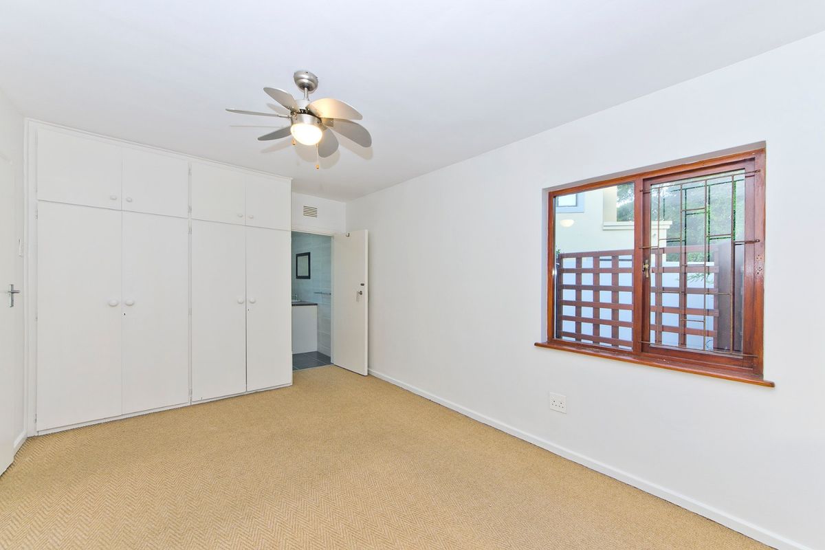 A bright, empty bedroom featuring built-in cupboards, a ceiling fan, and a timber-framed window with security bars.