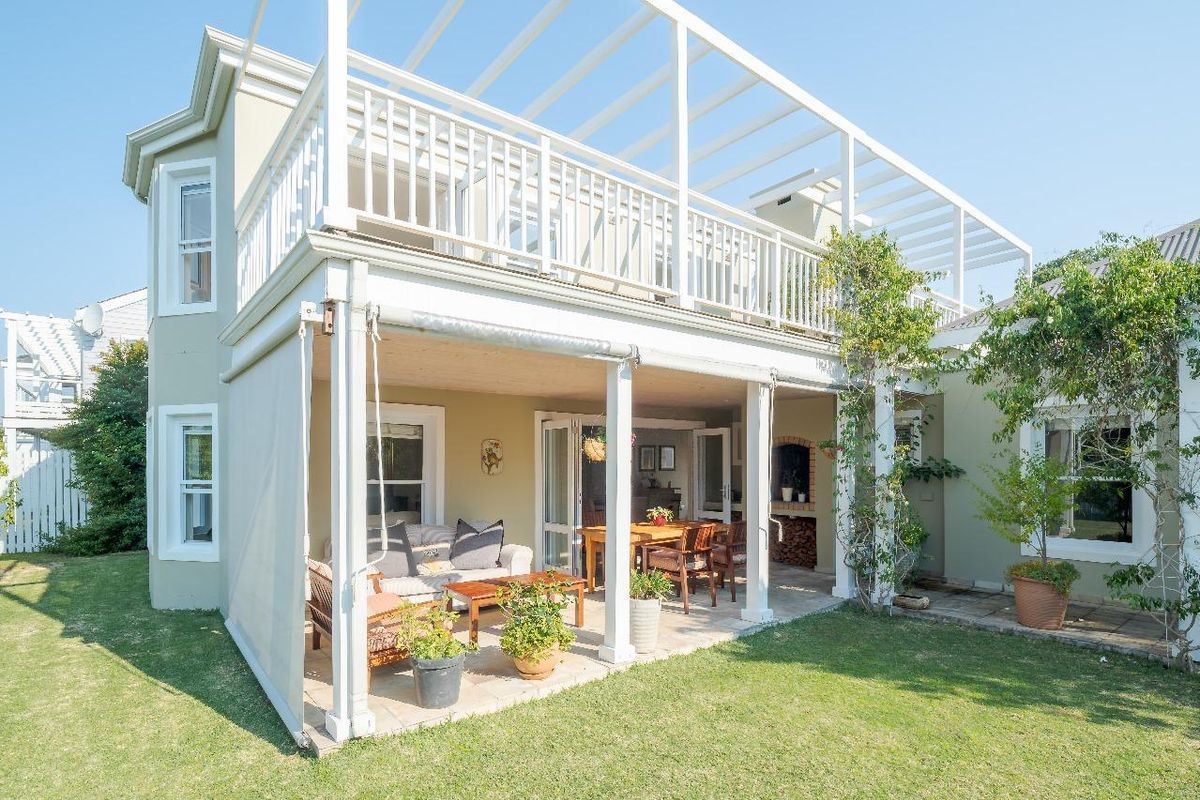 Rear view of the property featuring a covered patio with a built-in braai, leading to a spacious upper balcony with pergola structure.