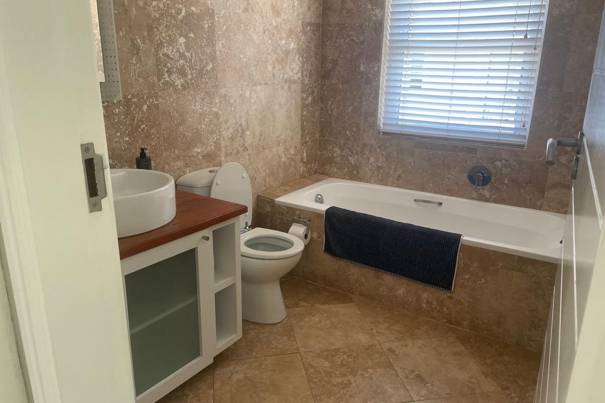 A well-maintained bathroom featuring travertine wall and floor tiling, a built-in bathtub, a modern vessel sink vanity, and a window with venetian blinds.