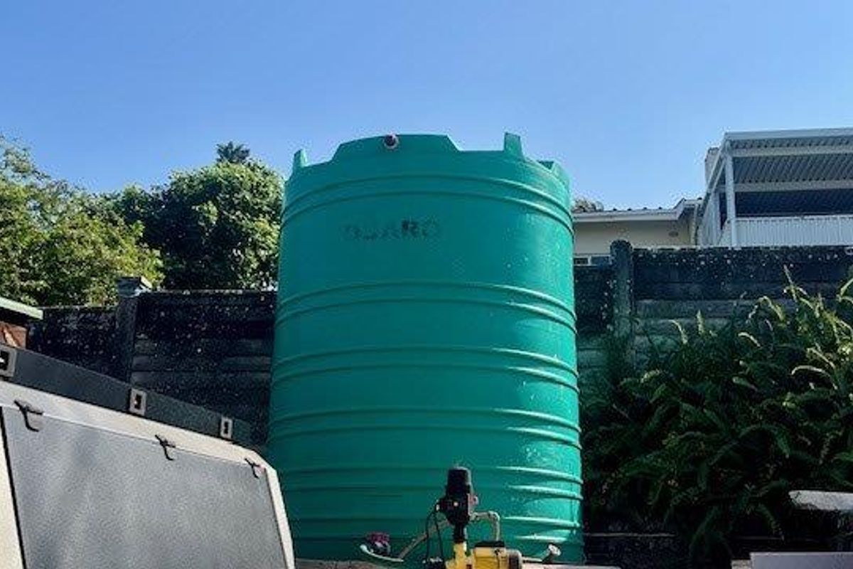 A large green water storage tank with an integrated pump system located in the rear garden area against a concrete boundary wall.