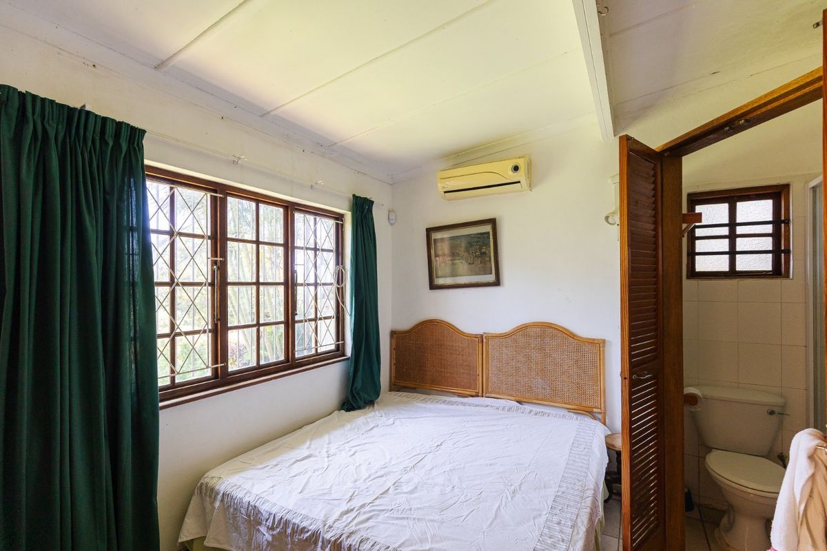 A compact bedroom featuring timber-framed windows with security bars, a wall-mounted air conditioner, and an attached en-suite bathroom with a louvered door.