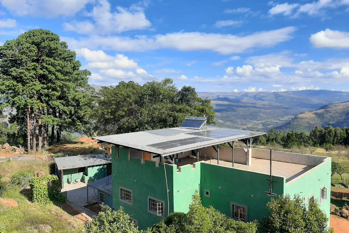 Aerial view of a green farmhouse featuring a flat roof terrace, solar energy installation, and expansive mountain landscape views.