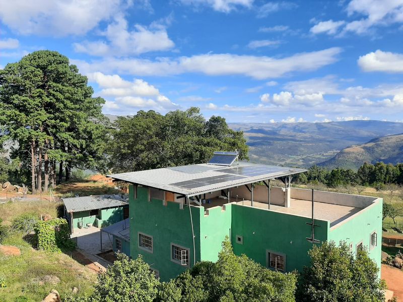 Aerial view of a green farmhouse featuring a flat roof terrace, solar energy installation, and expansive mountain landscape views.
