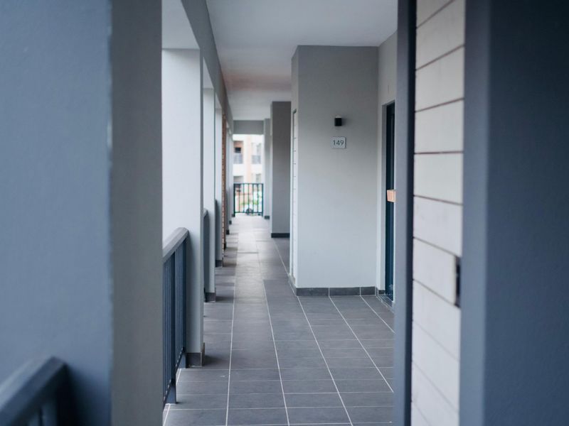 A clean, modern apartment hallway featuring grey tiled flooring, metal safety railings, and neutral wall finishes leading to residential unit entrances.