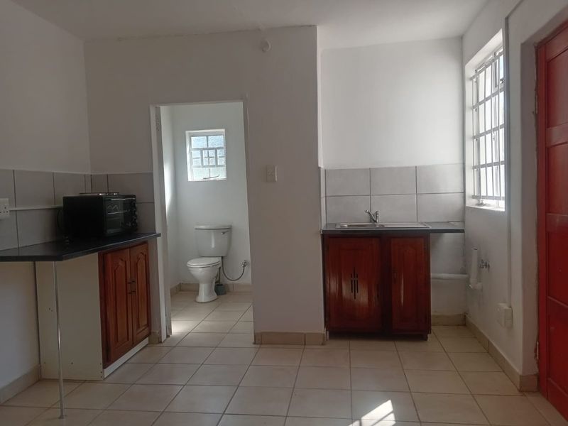Open plan kitchen area featuring wooden cabinetry, tiled splashbacks, and an adjoining bathroom with a toilet, finished with neutral floor tiles.