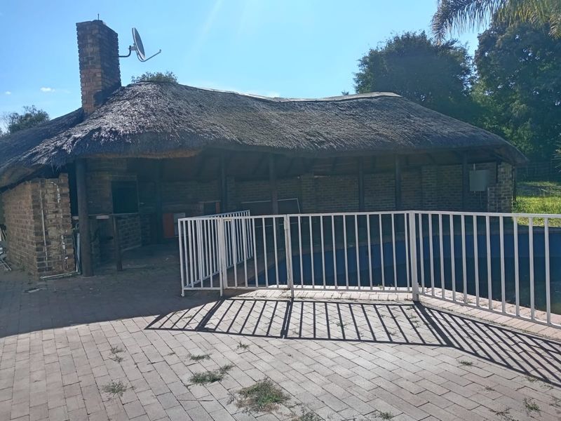 Outdoor pool area featuring a thatched lapa with a built-in braai, brick chimney, and paved surround, enclosed by a metal safety fence.