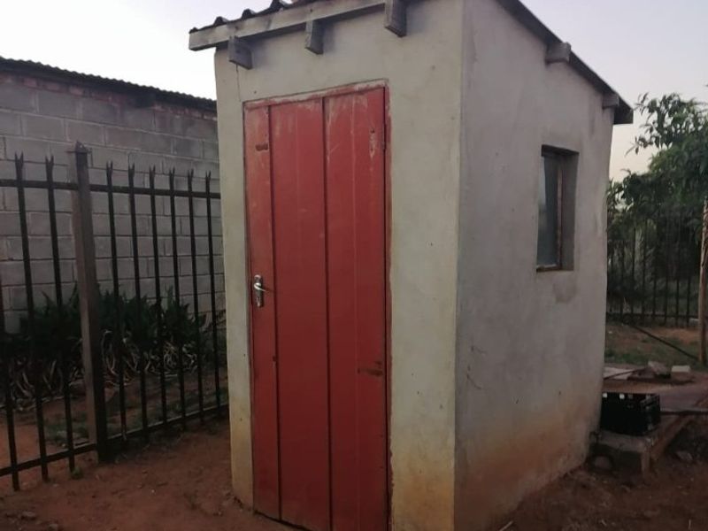 A small, detached utility structure featuring a red door, corrugated roof, and basic masonry construction, situated in a fenced outdoor area.