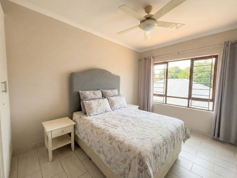 A well-maintained bedroom featuring neutral wall tones, tiled flooring, a ceiling fan, and a large window with security bars.