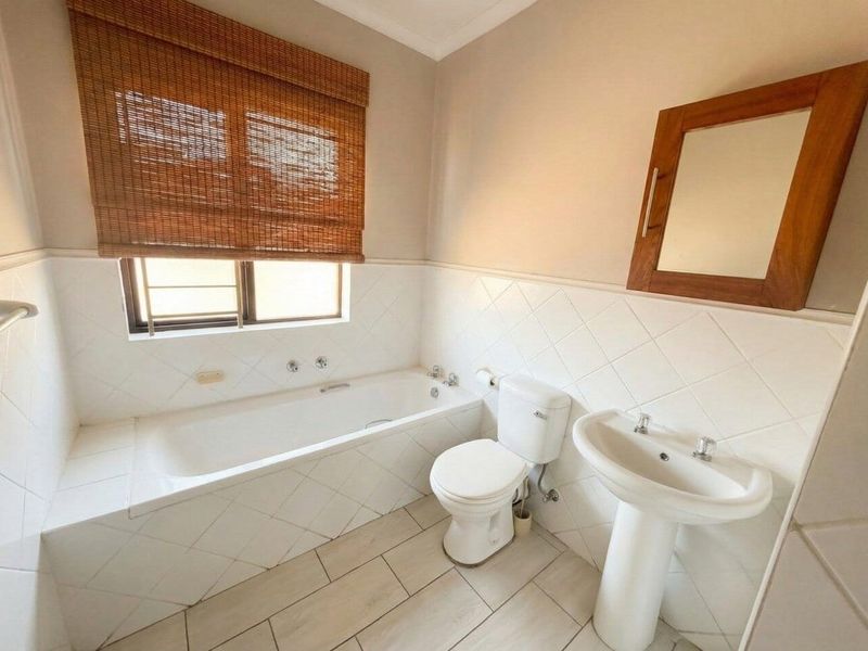 A functional bathroom featuring a built-in bathtub, pedestal sink, toilet, and neutral tiled finishes, offering a clean and practical space.