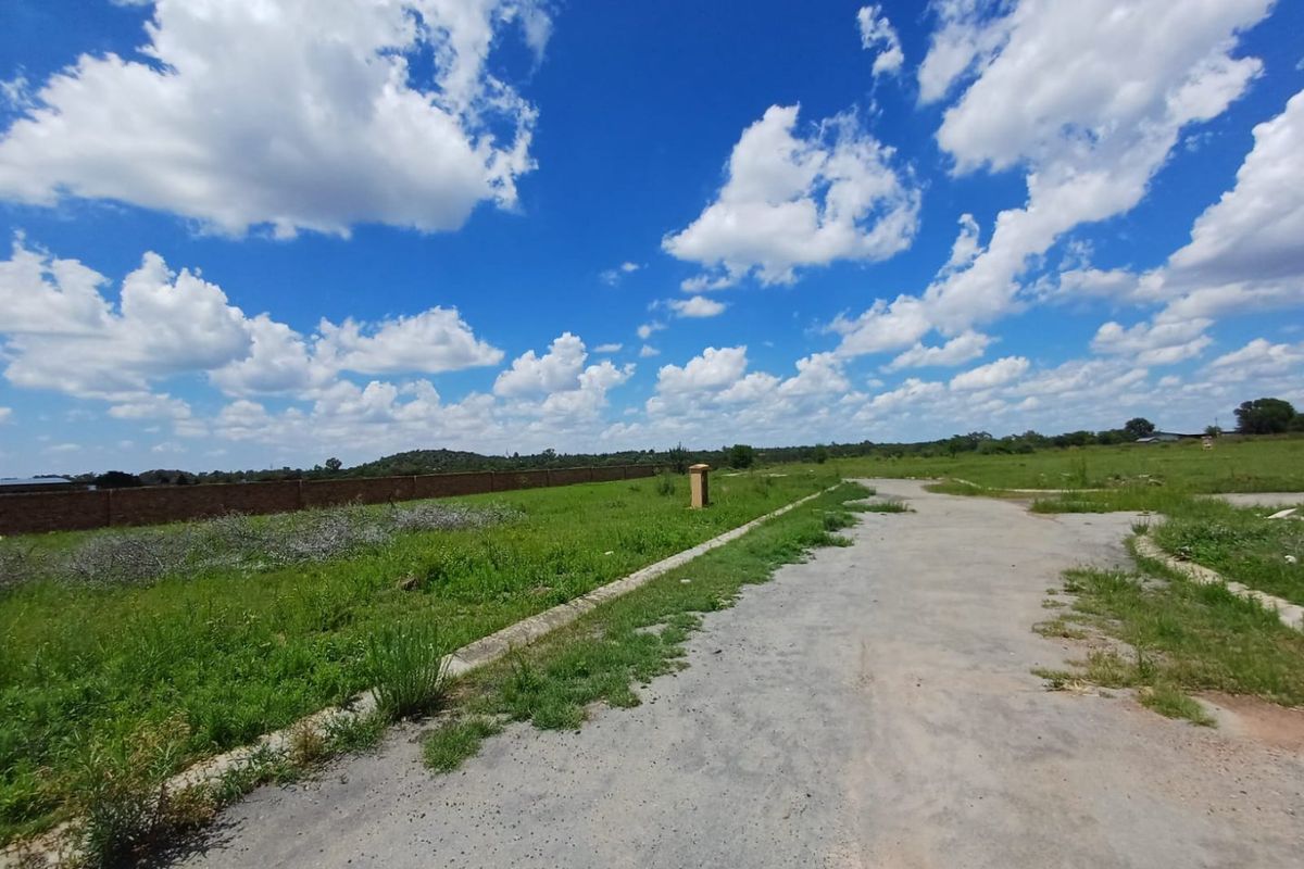 A view of a vacant residential plot featuring a paved access road, concrete curbing, and a brick boundary wall under a clear blue sky.