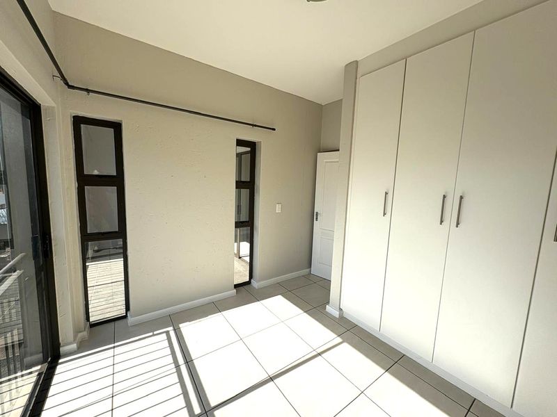 A bright, empty bedroom featuring neutral tiled flooring, built-in cupboards, and access to an outdoor area through a sliding glass door.