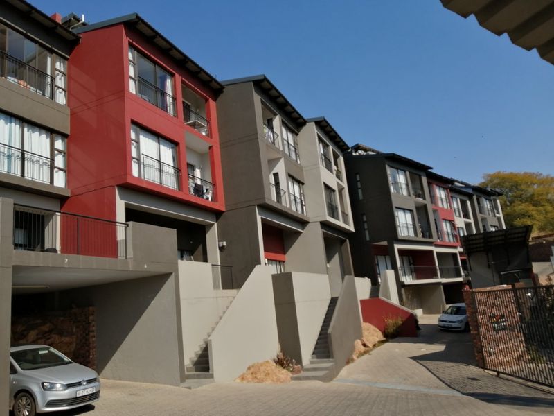 Modern multi-story apartment complex featuring a contemporary facade with red and grey accents, private balconies, and paved driveway access.