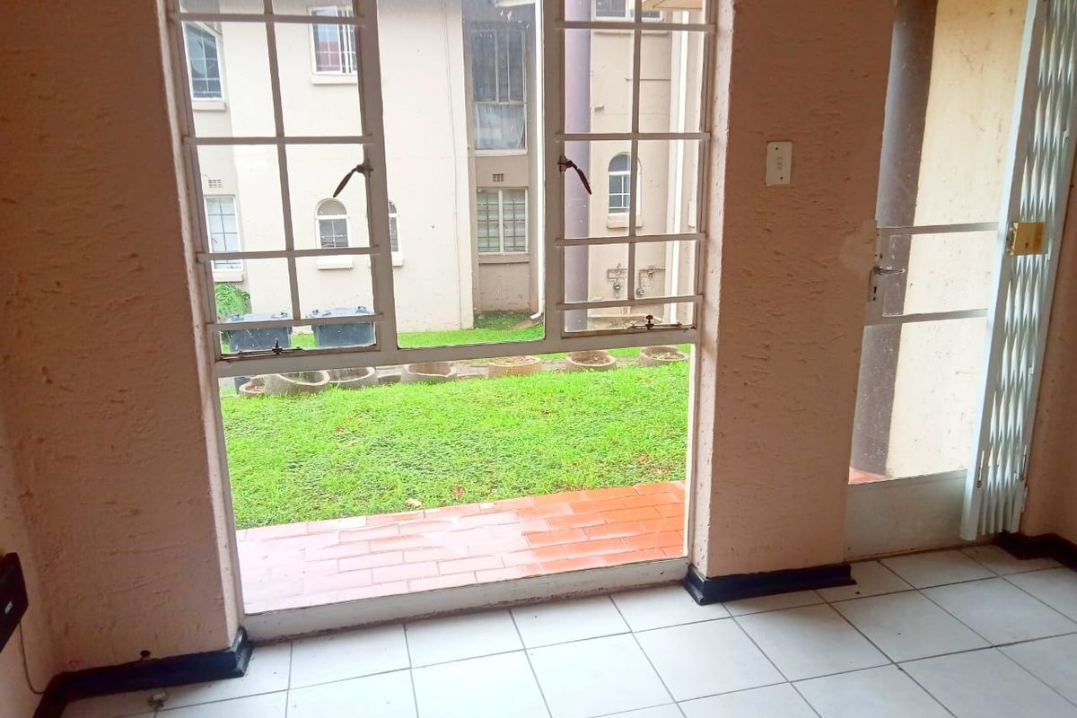 An interior view looking out through a large window and security gate towards a communal garden area with tiled flooring.