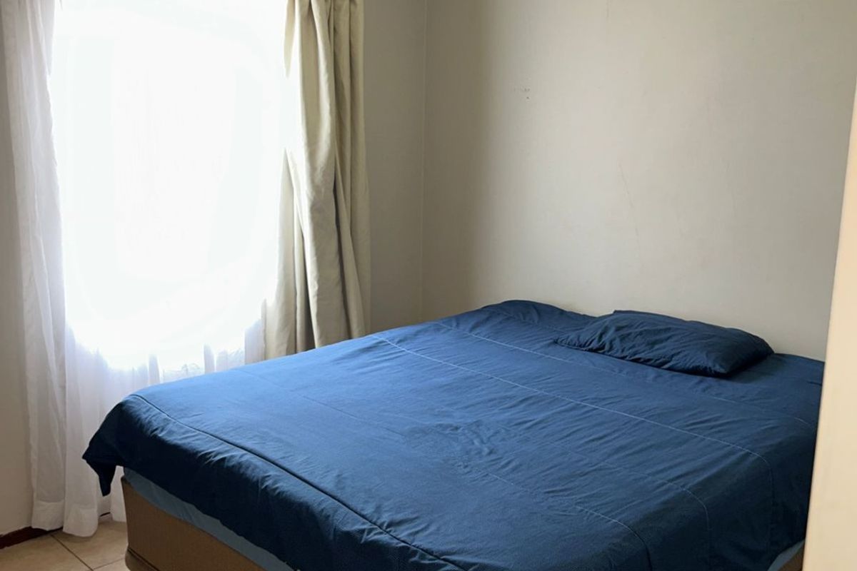 A simple bedroom featuring tiled flooring, a window with curtain tracks, and a wall-mounted ventilation vent.