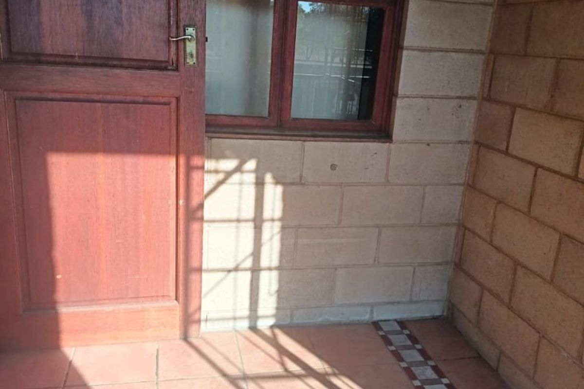 A covered patio area featuring tiled flooring, exposed brick walls, and a timber door with an adjacent window.
