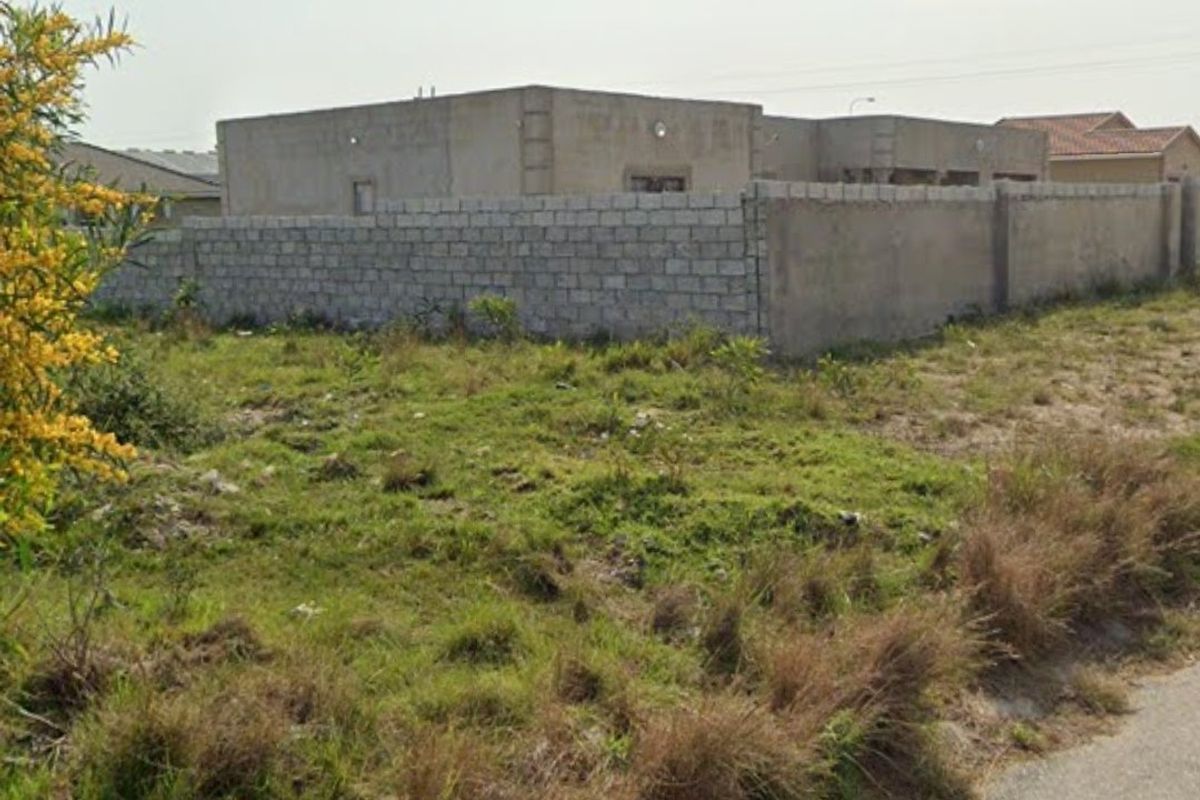 A vacant residential plot featuring natural grass cover and a perimeter concrete block wall, situated in a developed neighborhood.