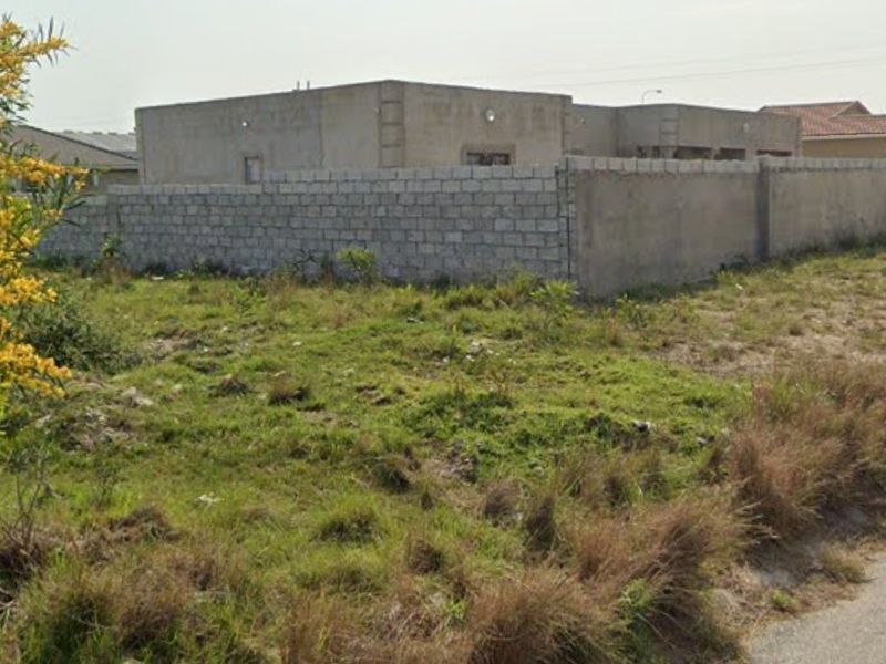 A vacant residential plot featuring natural grass cover and a perimeter concrete block wall, situated in a developed neighborhood.