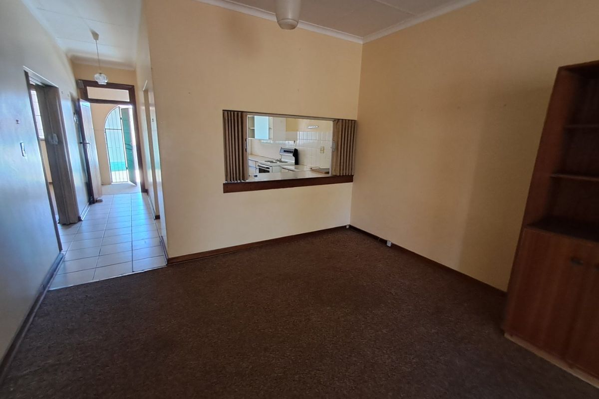 An empty living room featuring a serving hatch to the kitchen, tiled hallway access, and carpeted flooring, offering a blank canvas for renovation.