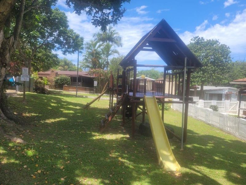 A communal garden area featuring a wooden playground structure with a slide, set within a landscaped lawn with mature trees and picket fencing.