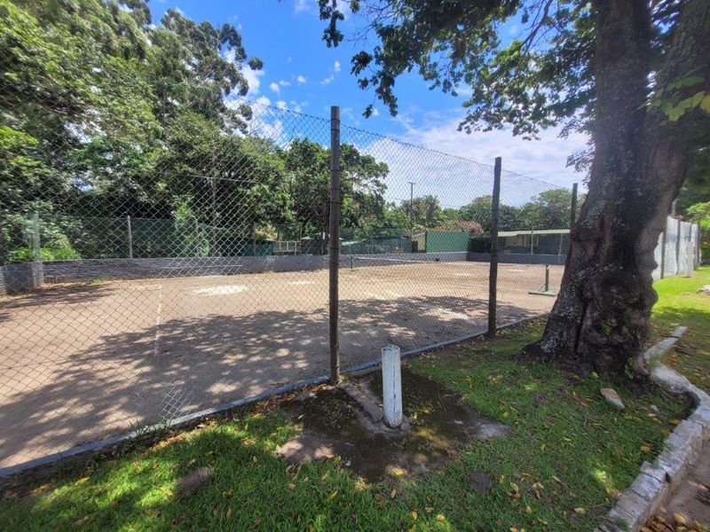 Outdoor tennis court facility featuring chain-link perimeter fencing and a paved playing surface, situated within a landscaped estate setting.